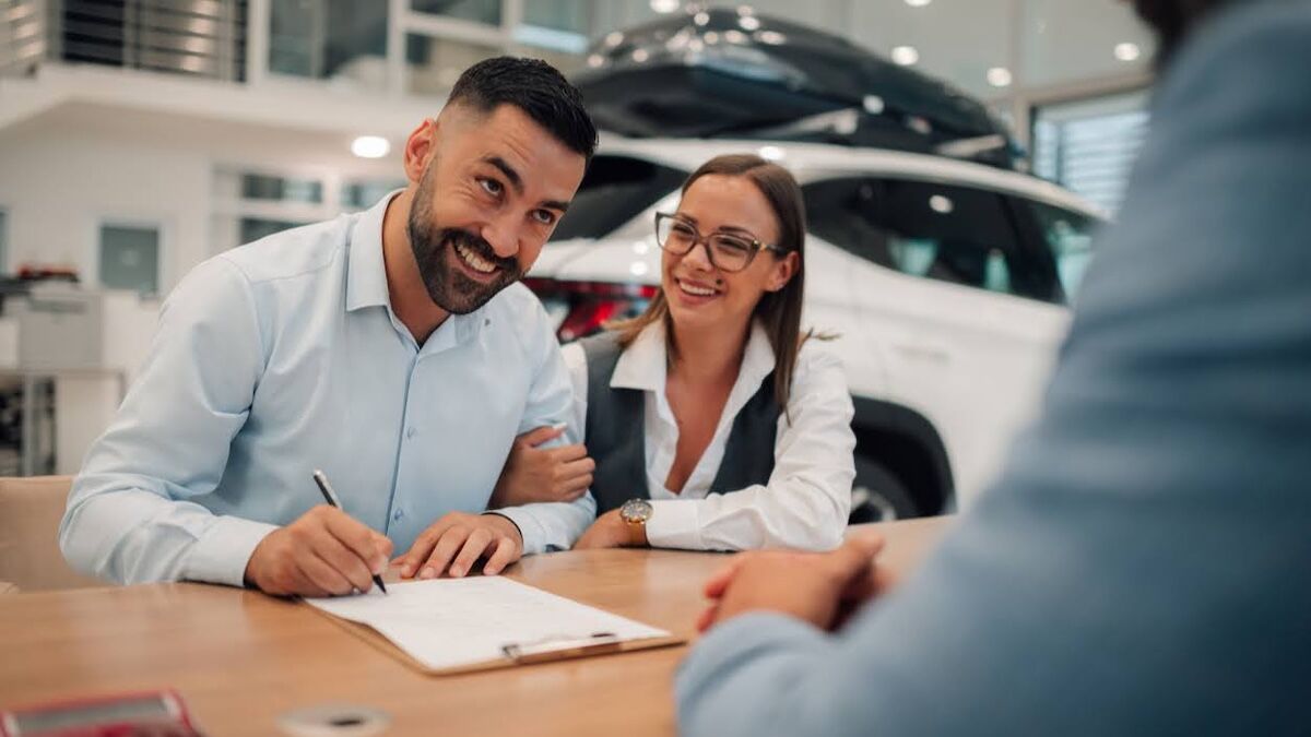 a couple smiles while signing paperwork in a car dealer's showroom